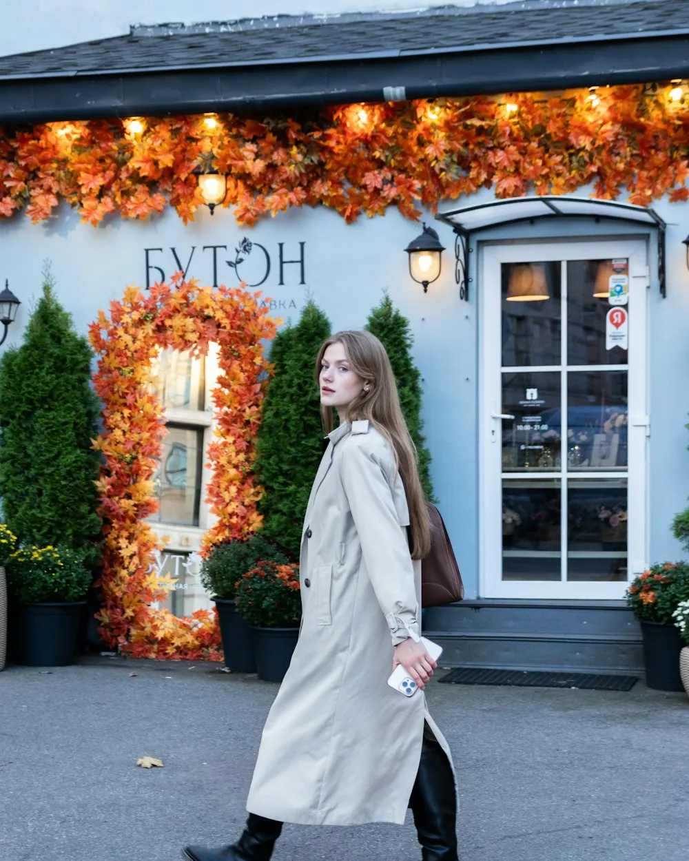 Woman in a beige trench coat walking past an autumn storefront