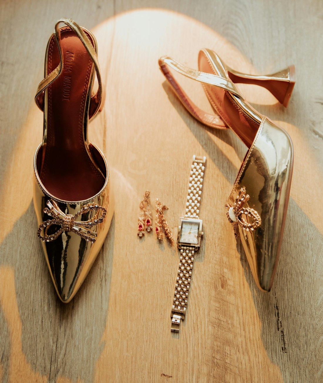 Flat lay of gold heels, a gold watch and drop earrings on a wooden surface