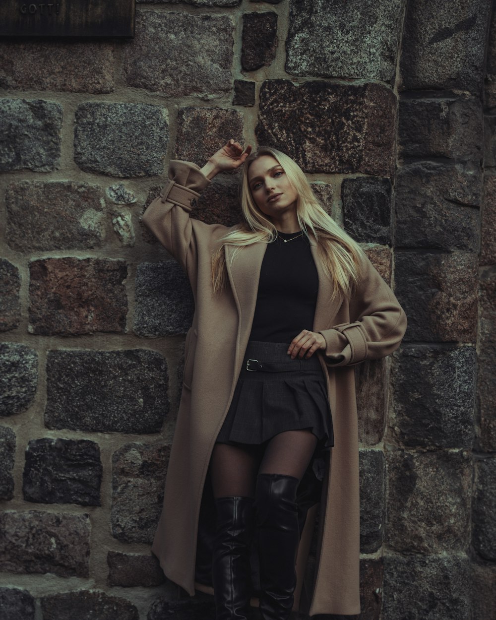 Blonde woman in a camel coat, pleated skirt and tall boots by a stone wall