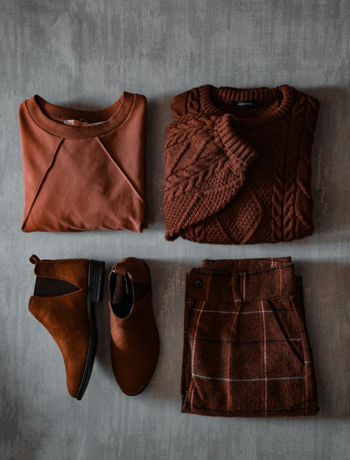 Flat lay of a rust cable knit, brown suede boots and a plaid skirt on a grey backdrop